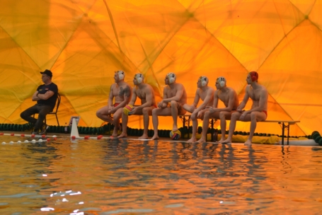 Sechs Wasserballspieler mit Badekappen und Badeanzügen sitzen auf einer Bank neben einem Schwimmbecken, an dessen Ende ein Trainer sitzt. Eine leuchtend gelbe, kuppelartige Struktur bildet den Hintergrund.