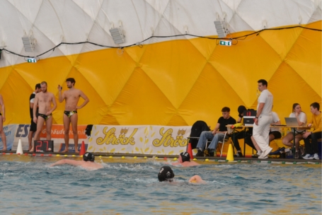 Wasserballspieler treten in einem Becken gegeneinander an, während Mannschaftskameraden und Offizielle an der Seite stehen und sitzen, vor einer gelben, geometrisch gemusterten Wand und einer Anzeigetafel mit 27 Punkten. Einige Spieler tragen schwarze Kappen mit Nummern.