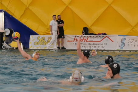 Ein Wasserballspieler mit einer gelben Mütze bereitet sich auf den Wurf vor, während seine Mannschaftskameraden und Gegner mit schwarzen Mützen im Becken verteidigen. Zwei Trainer stehen an einer gelben aufblasbaren Wand im Hintergrund.