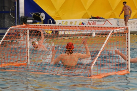 Ein Wasserballspiel, bei dem ein Spieler mit gelber Mütze versucht, ein Tor zu schießen, während der Torwart mit roter Mütze die Arme hebt, um den Schuss abzublocken. Das Spiel findet in einem Hallenbad mit einem leuchtend gelben Hintergrund statt.