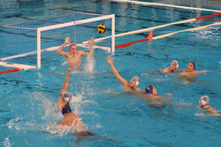 Ein laufendes Wasserballspiel mit Spielern, die blau-weiße Mützen tragen. Ein Spieler wirft den Ball in Richtung Tor, während der Torwart beide Hände hebt, um den Schuss abzuwehren. Andere Spieler schwimmen in der Nähe.
