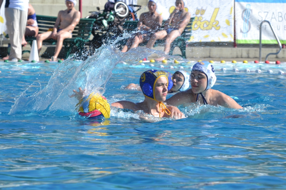 Zwei Wasserballspieler treten in einem Schwimmbecken gegeneinander an, wobei ein Spieler mit blauer Kappe den Ball hält, während er von einem Spieler mit weißer Kappe genau bewacht wird. Um sie herum sind Wasserspritzer zu sehen. Im Hintergrund sitzen Zuschauer.