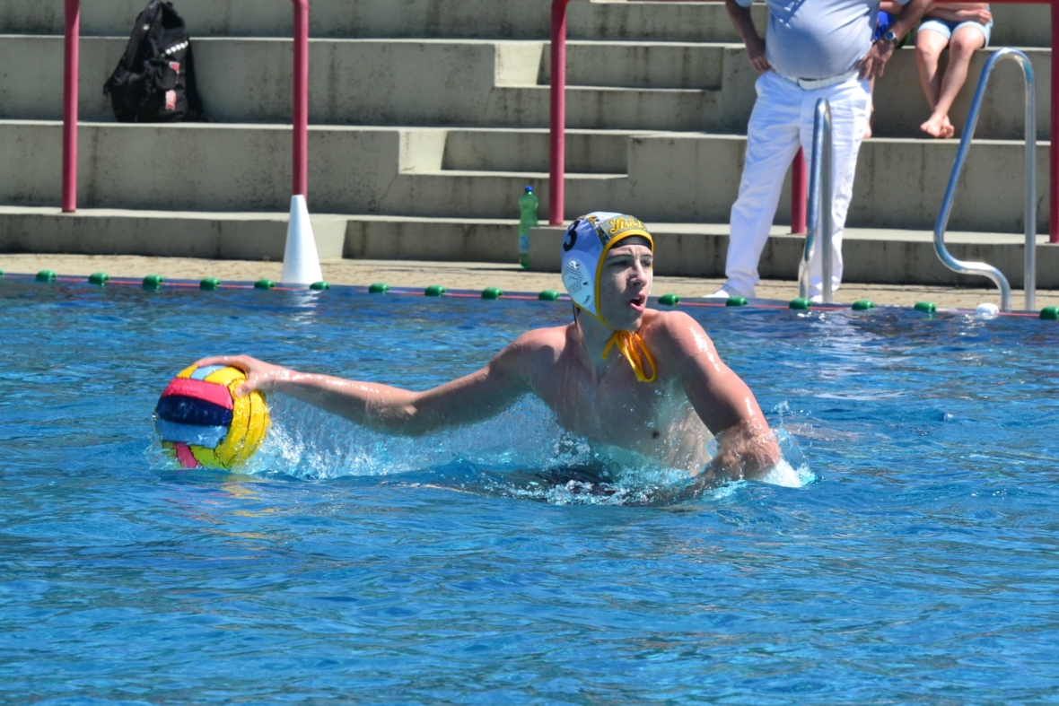Ein männlicher Wasserballspieler mit weißer Kappe bereitet sich darauf vor, einen bunten Ball in ein Schwimmbecken zu werfen, während im Hintergrund ein Zuschauer auf Stufen steht.