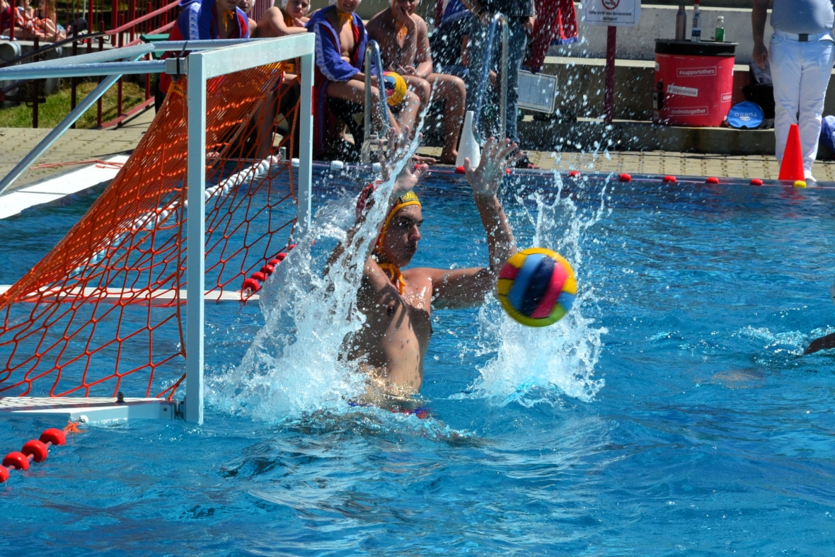 Ein Wasserballtorhüter blockiert einen bunten Ball, der in einem hellen Außenbecken spritzt, während die Zuschauer vom Beckenrand aus zusehen.