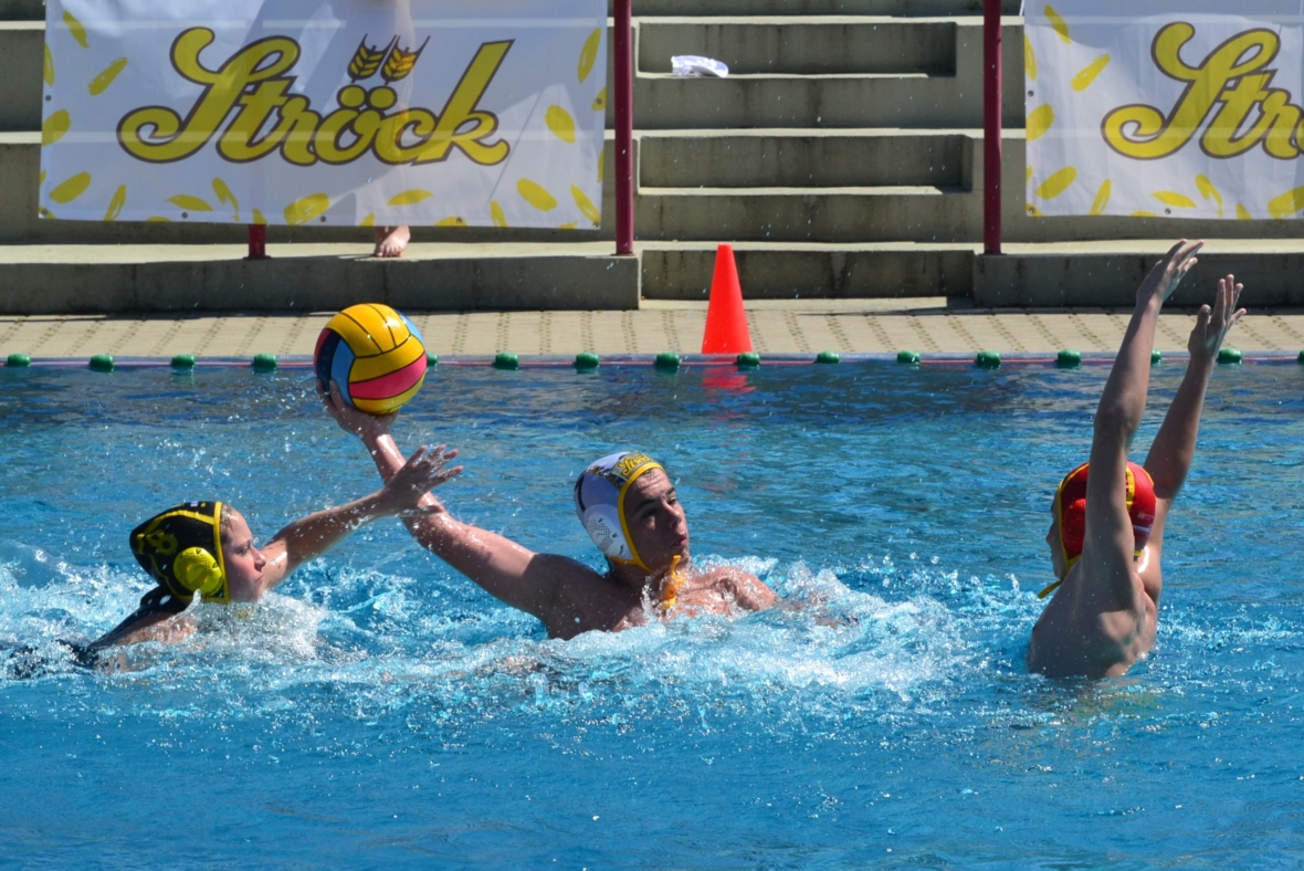 Drei Wasserballspieler treten in einem Schwimmbecken gegeneinander an. Ein Spieler in der Mitte bereitet sich darauf vor, den Ball zu werfen, während zwei Verteidiger ihre Arme zum Blocken heben. Im Hintergrund sind Stufen, Fahnen und ein Kegel zu sehen.