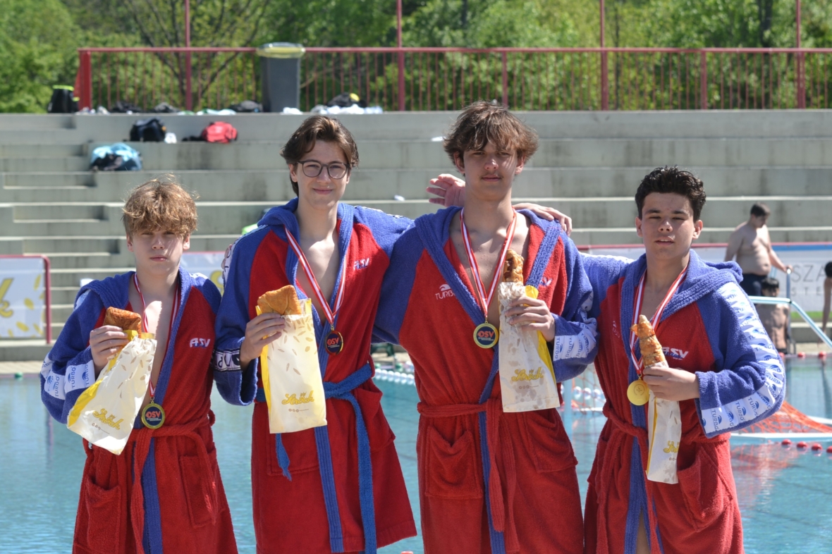 Vier junge männliche Schwimmer in roten und blauen Gewändern stehen nebeneinander an einem Schwimmbecken, jeder hält ein Baguette und eine Medaille und lächelt in die Kamera. Im Hintergrund sind Stufen und Bäume zu sehen.