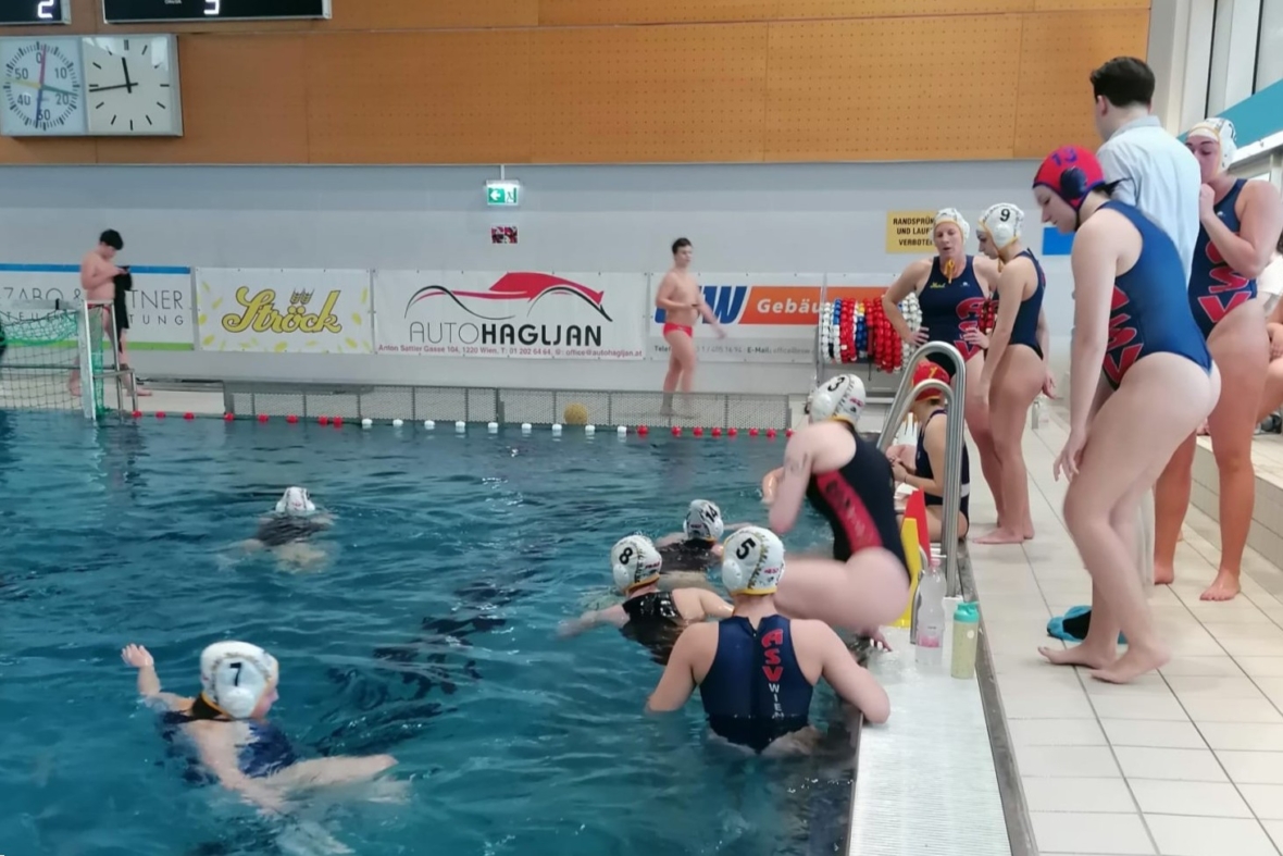 Eine Frauen-Wasserballmannschaft in Badeanzügen und Kappen versammelt sich am Beckenrand; einige Spielerinnen sind im Wasser, während andere am Beckenrand stehen oder sitzen, während ein Trainer in der Nähe Anweisungen gibt. Hallenbad mit sichtbaren Werbeschildern.