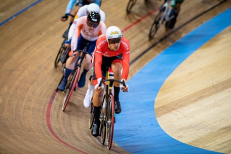 Eine Gruppe von Radfahrern fährt auf einer Indoor-Radrennbahn um die Wette. Der führende Fahrer trägt eine rote Uniform und einen weißen Helm, dicht gefolgt von anderen Fahrern in verschiedenfarbiger Kleidung.