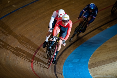 Drei Radfahrer fahren auf einer Indoor-Velodrombahn, tragen Helme und Fahrradkleidung. Der Spitzenreiter ist rot gekleidet, dicht gefolgt von den Teilnehmern in Weiß und Blau. Die Holzbahn ist mit roten und blauen Linien versehen.