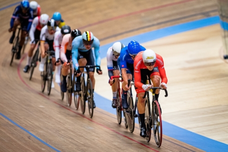 Eine Gruppe von Radrennfahrern, die eng beieinander auf einer Indoor-Radrennbahn fahren, wobei der führende Fahrer einen roten Anzug und einen Helm trägt, gefolgt von Teilnehmern in blauen und weißen Outfits.