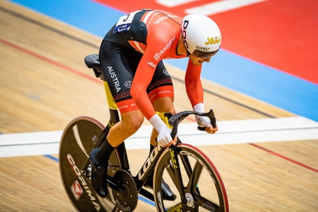Ein Radsportler in einem rot-schwarzen Austria-Team-Trikot und weißem Helm fährt auf einem schwarzen Bahnrad in einer Halle mit Holzboden und bunten Bahnlinien.