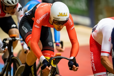 Ein Radfahrer in einer rot-schwarzen Uniform und mit weißem Helm fährt auf einer Indoor-Bahn, wobei er sich stark nach vorne lehnt und andere Fahrer in der Nähe zu sehen sind. Auf dem Trikot sind Österreich und mehrere Sponsorenlogos zu sehen.