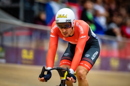 Ein Radfahrer in einer rot-schwarzen Uniform und einem weißen Helm fährt auf einem Indoor-Track-Bike. Auf der Uniform steht "Österreich", und der Radfahrer wirkt konzentriert, während das Publikum im Hintergrund verschwommen ist.