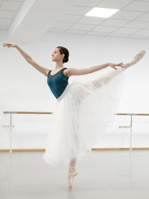 Eine Balletttänzerin in einem blaugrünen Trikot und einem weißen Tutu führt in einem hellen Tanzstudio mit verspiegelten Wänden und Ballettstangen eine Arabeske auf Spitzen vor.