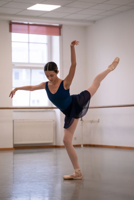 Eine Balletttänzerin in einem blauen Trikot und einem durchsichtigen Rock balanciert auf einem spitzen Fuß in einem Studio und hebt das andere Bein und einen Arm hoch, wobei sie unter dem natürlichen Licht der großen Fenster Anmut und Ausgeglichenheit zeigt.