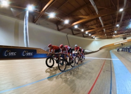 Vier rot-weiß gekleidete Radsportler, die eng beieinander auf einer hölzernen Radrennbahn unter einer hohen, beleuchteten Holzdecke und mit UEC-Bannern an den Wänden fahren.