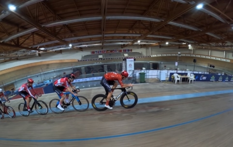 Drei Radsportler in roten und blauen Trikots rasen eng beieinander auf einer Indoor-Velodrombahn, im Hintergrund sind Holzbalken und leere Zuschauerplätze zu sehen.