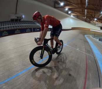 Ein Radfahrer mit rotem Helm und Trikot fährt mit einem Rennrad auf einer Indoor-Velodrombahn. Die Tribünen sind leer und die gebogene Holzbahn ist mit farbigen Linien umrandet.