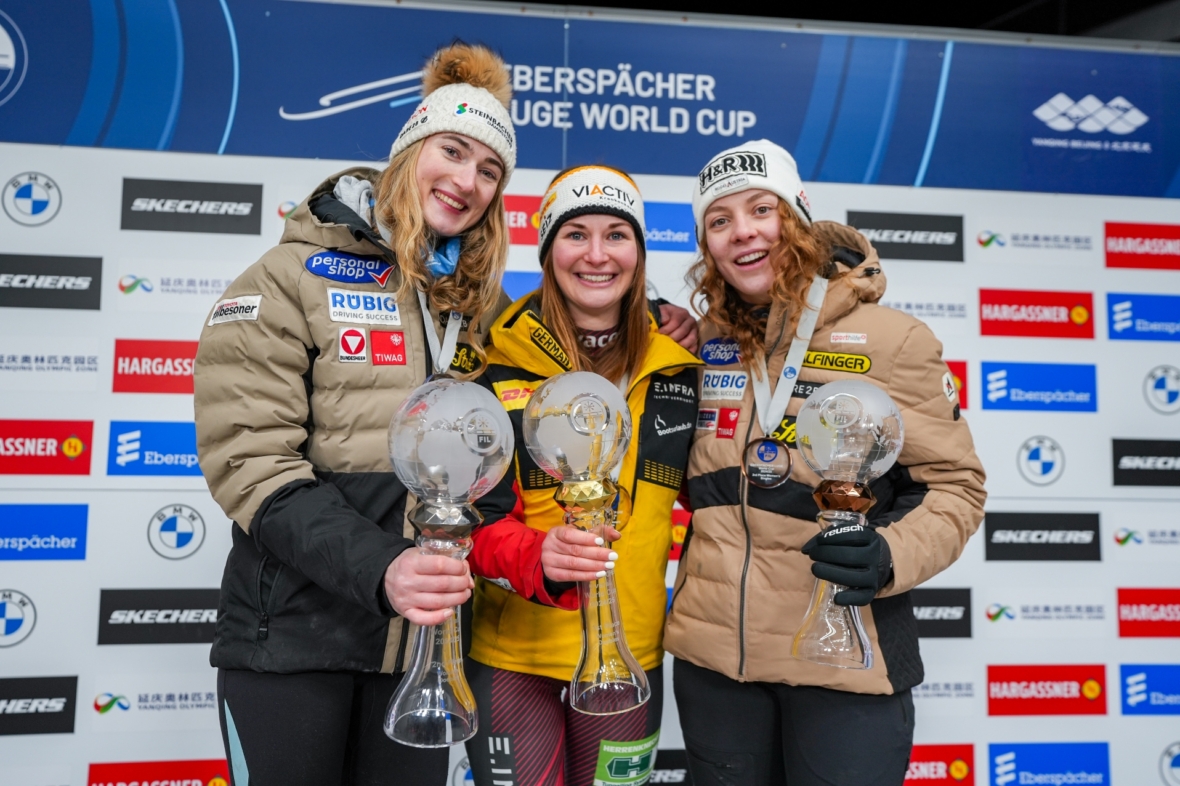 Drei Sportlerinnen in Winterjacken stehen lächelnd auf einem Podium, jede hält eine große Glastrophäe in der Hand, im Hintergrund sind die Logos der Sponsoren einer Sportveranstaltung zu sehen.