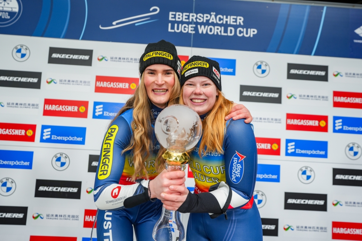 Zwei Sportlerinnen in blauen Anzügen stehen lächelnd mit einem großen Pokal in der Hand vor einer Kulisse mit Eberspächer Rennrodel-Weltcup und verschiedenen Sponsorenlogos.