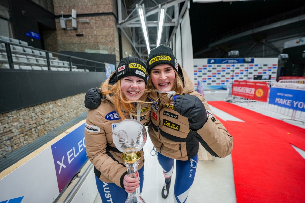 Zwei lächelnde Sportlerinnen in Wintersportkleidung posieren zusammen, eine hält eine große Meisterschaftstrophäe. Sie stehen auf einem roten Teppich mit Veranstaltungsbannern und einem Stadion im Hintergrund.