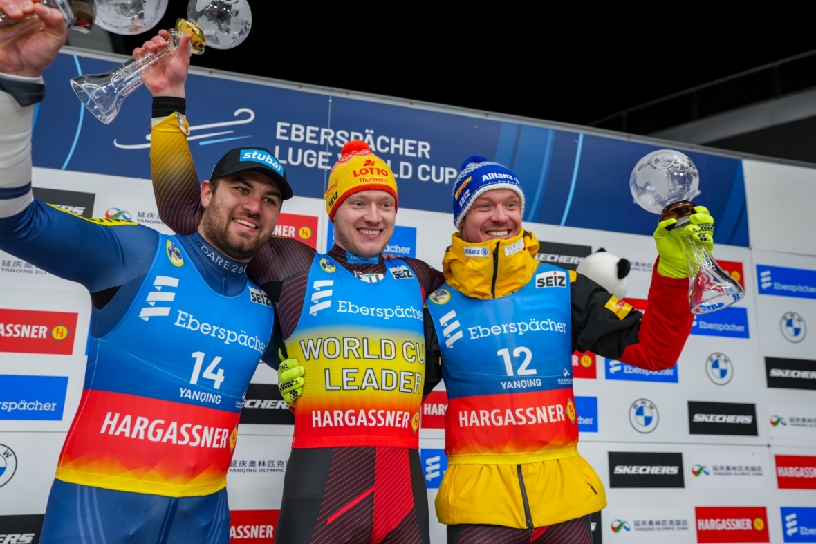 Drei männliche Rennrodler in Rennanzügen und Helmen stehen lächelnd auf einem Podium und halten Glastrophäen in der Hand. Im Hintergrund sind der Eberspächer Rennrodel-Weltcup und verschiedene Sponsorenlogos zu sehen.