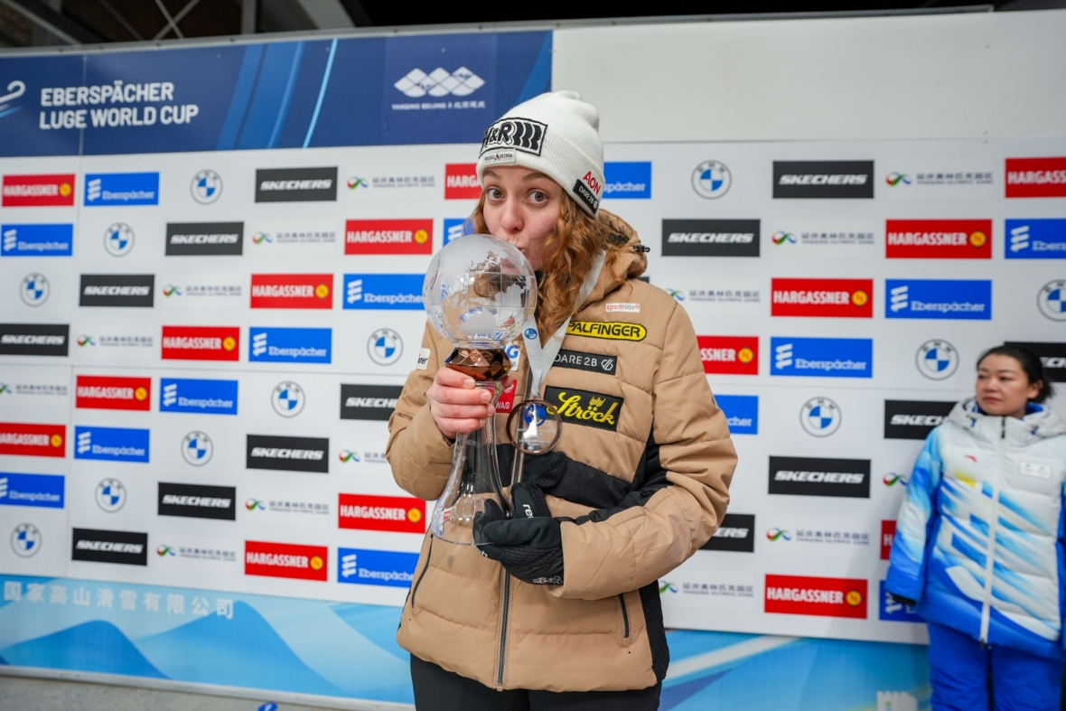 Eine Sportlerin in hellbrauner Jacke und weißer Mütze küsst eine große Glastrophäe auf einem Podium beim Eberspächer Rennrodel-Weltcup. Im Hintergrund sind Sponsorenlogos und eine weitere Person zu sehen.