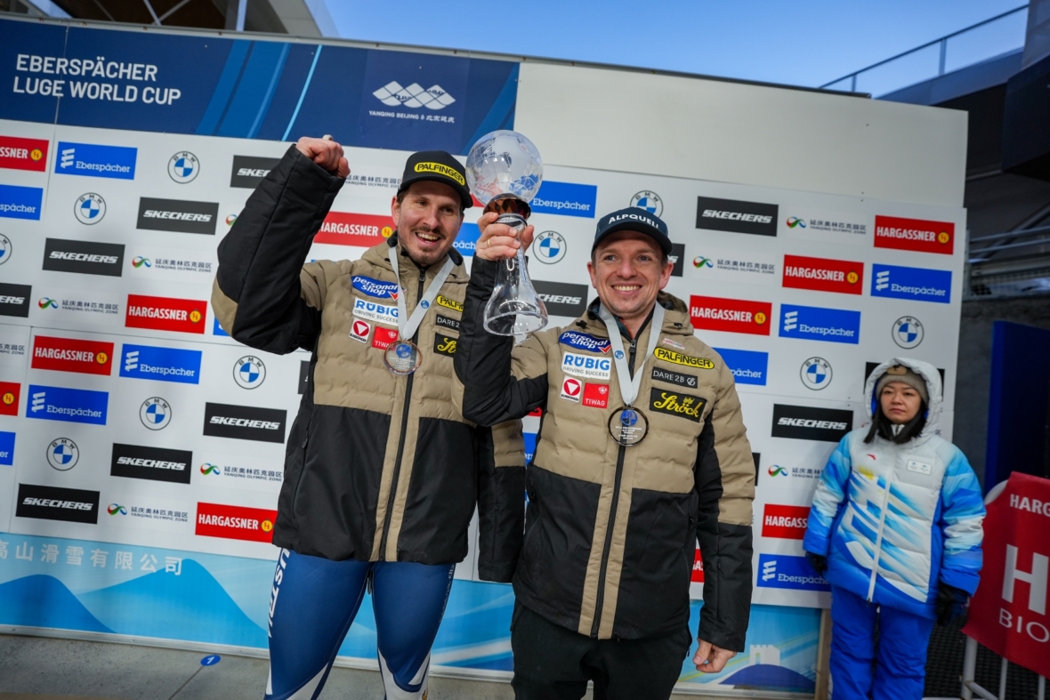 Zwei männliche Athleten in Wintersportkleidung stehen lächelnd auf einem Podium, einer hält eine große Glastrophäe. Beide tragen Medaillen und gebrandete Jacken. Im Hintergrund sind Veranstaltungsschilder und eine Person in einem blauen Mantel zu sehen.