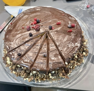 Ein Schokoladenkuchen mit glänzender Schokoladenglasur, in Scheiben geschnitten, mit verschiedenen Beeren belegt und mit einer Krümelmischung um den Boden dekoriert, serviert auf einer Glasplatte.