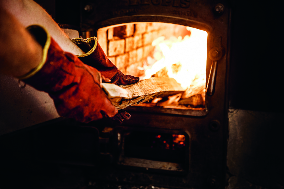 Eine Person mit roten Schutzhandschuhen legt ein Stück Holz in einen brennenden Ofen, wobei im Inneren des Metallofens helle Flammen zu sehen sind.