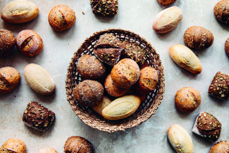 Ein geflochtener Korb, gefüllt mit verschiedenen Brötchen, steht auf einer hellen Fläche, umgeben von weiteren runden und ovalen Brötchen, die um ihn herum verteilt sind.