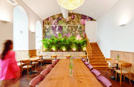 Ein heller, moderner Essbereich mit einem langen Holztisch, hellen Holzstühlen, üppigen grünen Pflanzen, einem Blumenwandbild, Bogenfenstern, einem Kronleuchter und einer Treppe. Eine Person in Rosa geht an den Tischen vorbei.