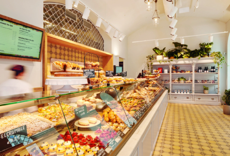 Ein heller Bäckerei-Innenraum mit einer Glasauslage, gefüllt mit verschiedenen Backwaren, Kuchen und Desserts. Regale mit Pflanzen und Produkten säumen die Rückwand, und links hängt eine digitale Speisekarte. Der Boden hat ein gelbes, geometrisches Muster.