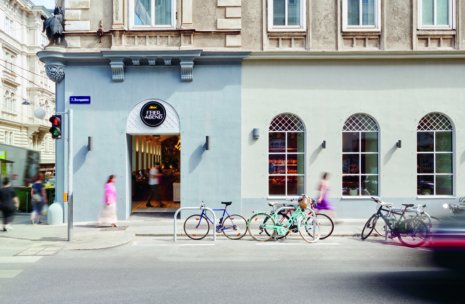 Eine Straßenansicht eines Café-Eingangs mit dem Schild FEIERABEND. Mehrere Fahrräder sind draußen geparkt, und Menschen gehen an dem hellblaugrauen Gebäude mit gewölbten Fenstern vorbei. Der Verkehr und die unscharfen Bewegungen deuten auf ein geschäftiges städtisches Umfeld hin.