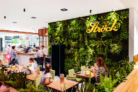 Ein modernes Café mit einer grünen Pflanzenwand, dem leuchtend gelben Schild "Ströck", Holztischen, grünen Stühlen und Menschen, die dort essen. Der Raum hat ein natürliches, frisches Ambiente mit viel Grün.