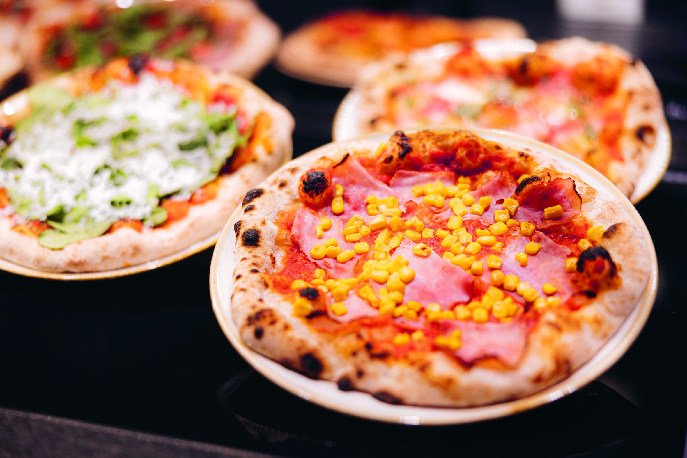 Nahaufnahme mehrerer Pizzen auf Tellern mit einer mit Schinken und Mais belegten Pizza im Vordergrund und anderen Pizzen mit grünem Blattbelag und Käse im Hintergrund auf einer schwarzen Fläche.