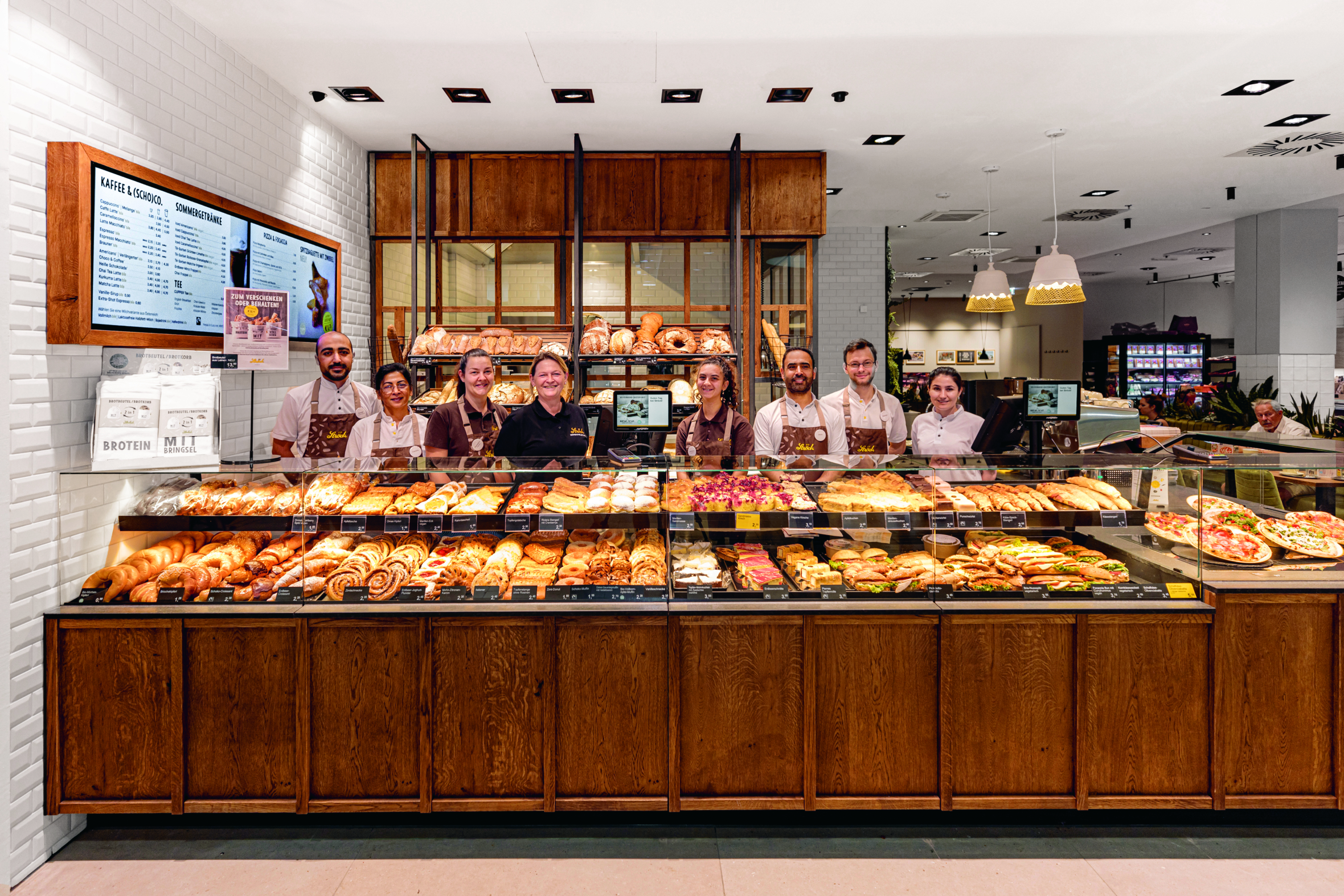 Eine Gruppe von Bäckereimitarbeitern steht in einer hellen, modernen Bäckerei mit Holztheke hinter einer Glasauslage mit verschiedenen Brotsorten, Gebäck und Backwaren und lächelt in die Kamera.