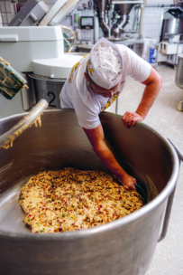 Ein Bäcker in weißer Uniform und mit Hut mischt eine große Menge Teig mit bunten Zutaten in einem großen industriellen Metallmixer in einer Bäckerei.