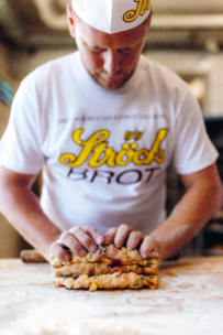 Ein Bäcker, der ein weißes Hemd und eine Mütze mit gelbem Ströck BROT-Schriftzug trägt, formt mit beiden Händen Teig auf einer bemehlten Fläche und konzentriert sich auf seine Arbeit.