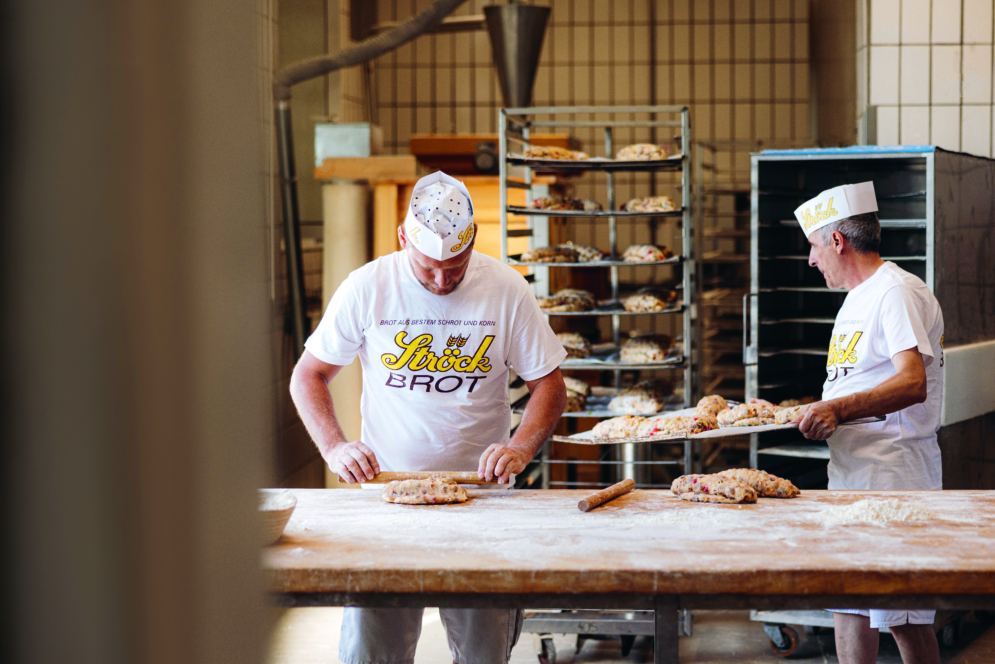 Zwei Bäcker in weißen Uniformen und mit Hüten bereiten den Teig auf einem bemehlten Holztisch in einer Bäckerei zu. Hinter ihnen befinden sich Regale mit Teig, im Hintergrund sind Kachelwände zu sehen.