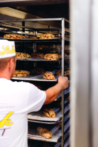 Eine Person in weißem Hemd und Hut schiebt ein großes Metallgestell mit Tabletts mit frisch gebackenem Gebäck in einen Ofen oder eine Bäckerei. Das Gebäck ist goldbraun und hat sichtbare Fruchtstücke.