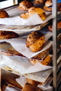 Goldbraune Brötchen mit Körnern kühlen auf mit Pergament ausgelegten Metallgestellen in einer Bäckerei ab. Die Gestelle sind vertikal gestapelt und das Brot sieht frisch gebacken und rustikal aus.