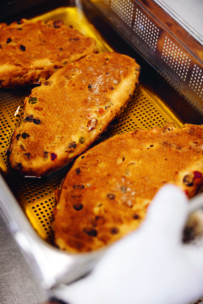 Drei Stücke goldbraunes, mit Früchten gefülltes Brot liegen auf einem perforierten Tablett, das wahrscheinlich gerade gebacken wurde. In der unteren rechten Ecke ist ein kleiner Teil eines weißen Tuches oder Handschuhs zu sehen.