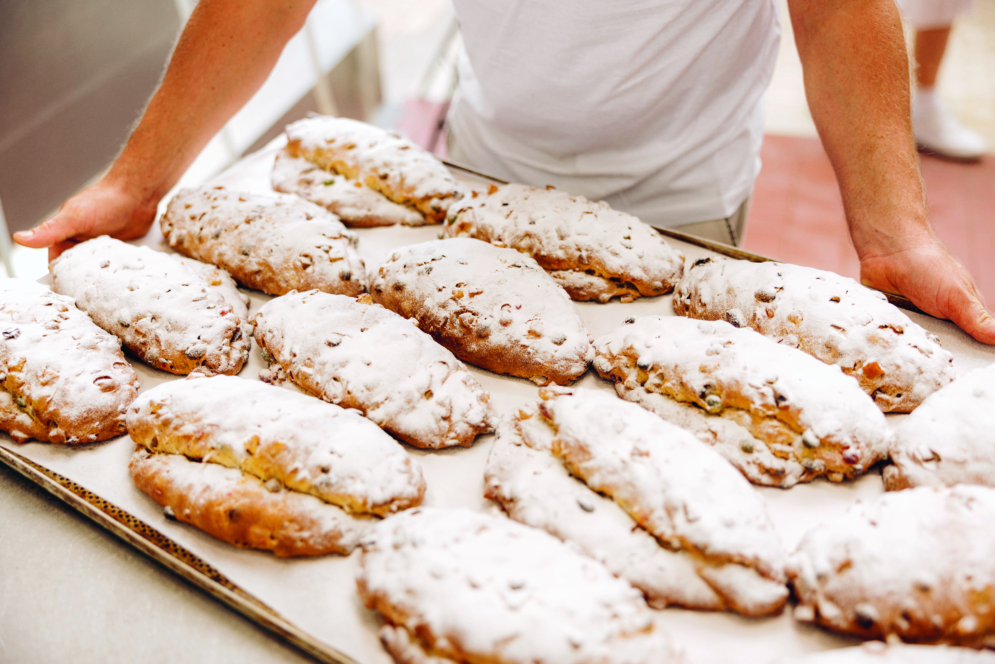 Eine Person hält ein großes Tablett mit frisch gebackenem, mit Puderzucker bestreutem Gebäck, möglicherweise deutscher Stollen, der in Reihen auf Pergamentpapier angeordnet ist.