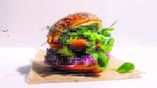 Ein Gourmet-Veggie-Burger mit entkerntem Brötchen, Blattgemüse, violettem Kohl, knusprig gebratenem Patty und cremiger Soße liegt auf braunem Pergamentpapier vor einem hellen, minimalen Hintergrund.