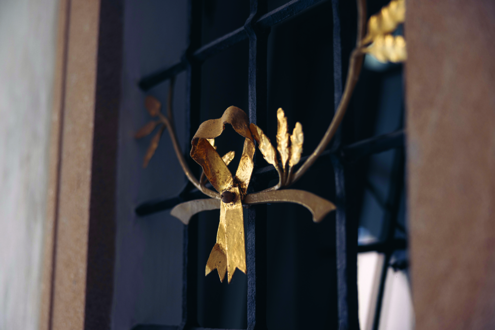 Nahaufnahme eines verzierten Fenstergitters mit einem dekorativen Element aus goldfarbenen Metallblättern und -bändern, das auf einem schwarzen Gitter vor einem dunklen Hintergrund angebracht ist.