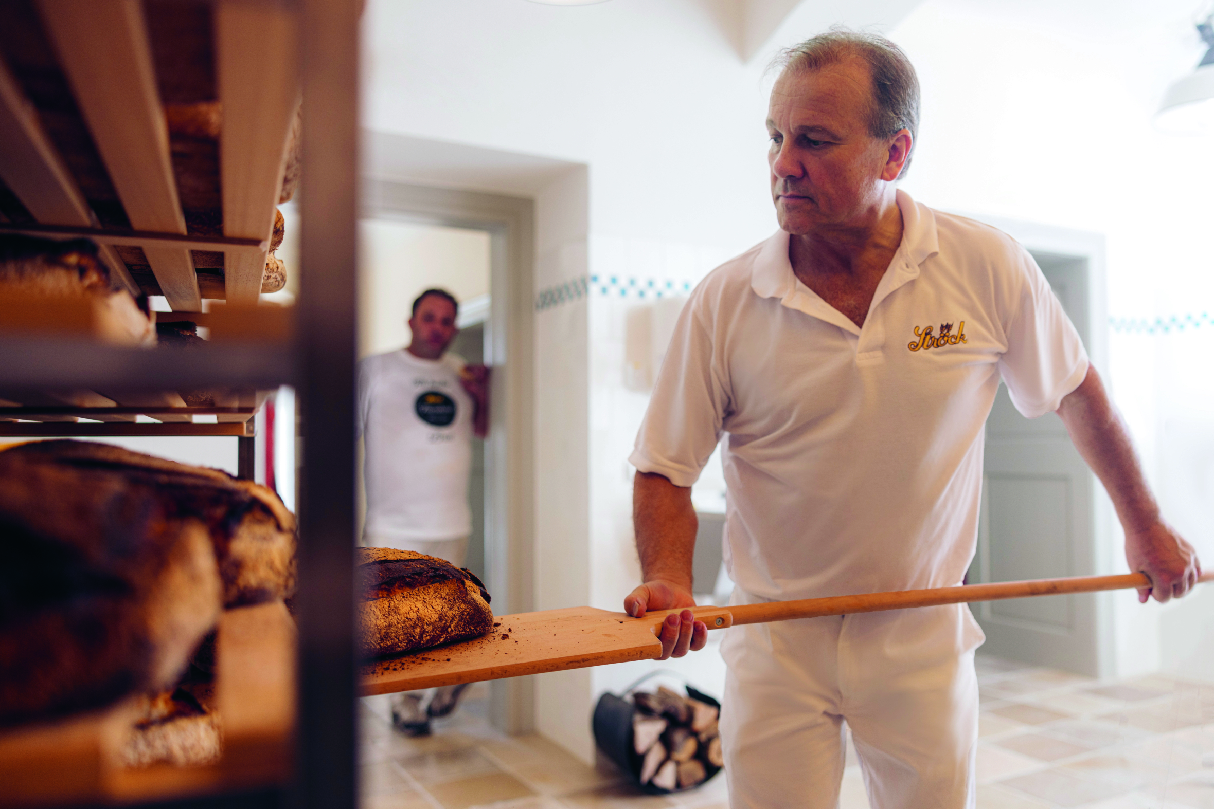 Ein Bäcker in weißer Uniform nimmt mit einer hölzernen Schale frisch gebackenes Brot aus einem Regal, während eine andere Person im Hintergrund in der Nähe einer Tür in einer hellen Bäckerei steht.