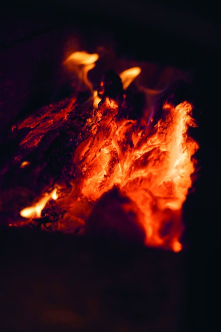 Nahaufnahme von rot und orange glühender Glut und brennendem Holz, mit hellen Flammen und verkohlten Strukturen, die ein warmes und intensives Feuer erzeugen.