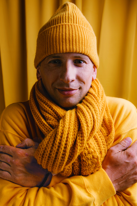 Ein Mann, der eine gelbe Strickmütze, einen dicken gelben Schal und einen gelben Pullover trägt, steht mit verschränkten Armen und lächelt leicht. Der Hintergrund ist ebenfalls gelb, wodurch ein monochromer Look entsteht.