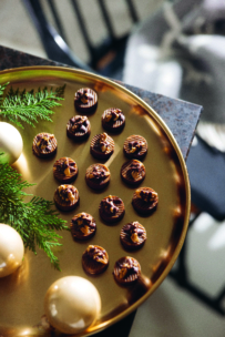 Ein goldenes Tablett mit kleinen, dekorativen Schokoladenpralinen, die in ordentlichen Reihen angeordnet sind, neben grünen, immergrünen Zweigen und großen, runden weißen Pralinen. Das Tablett steht auf einer dunklen Fläche mit einem unscharfen Hintergrund.